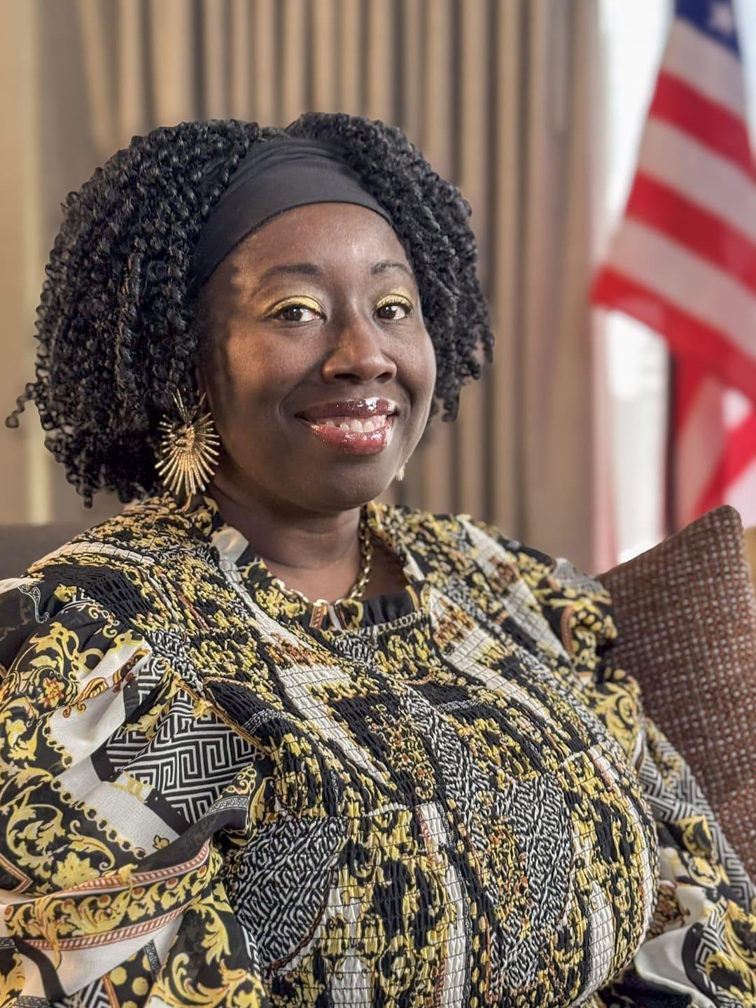 A woman wearing a patterned top sits indoors, looking toward the camera.