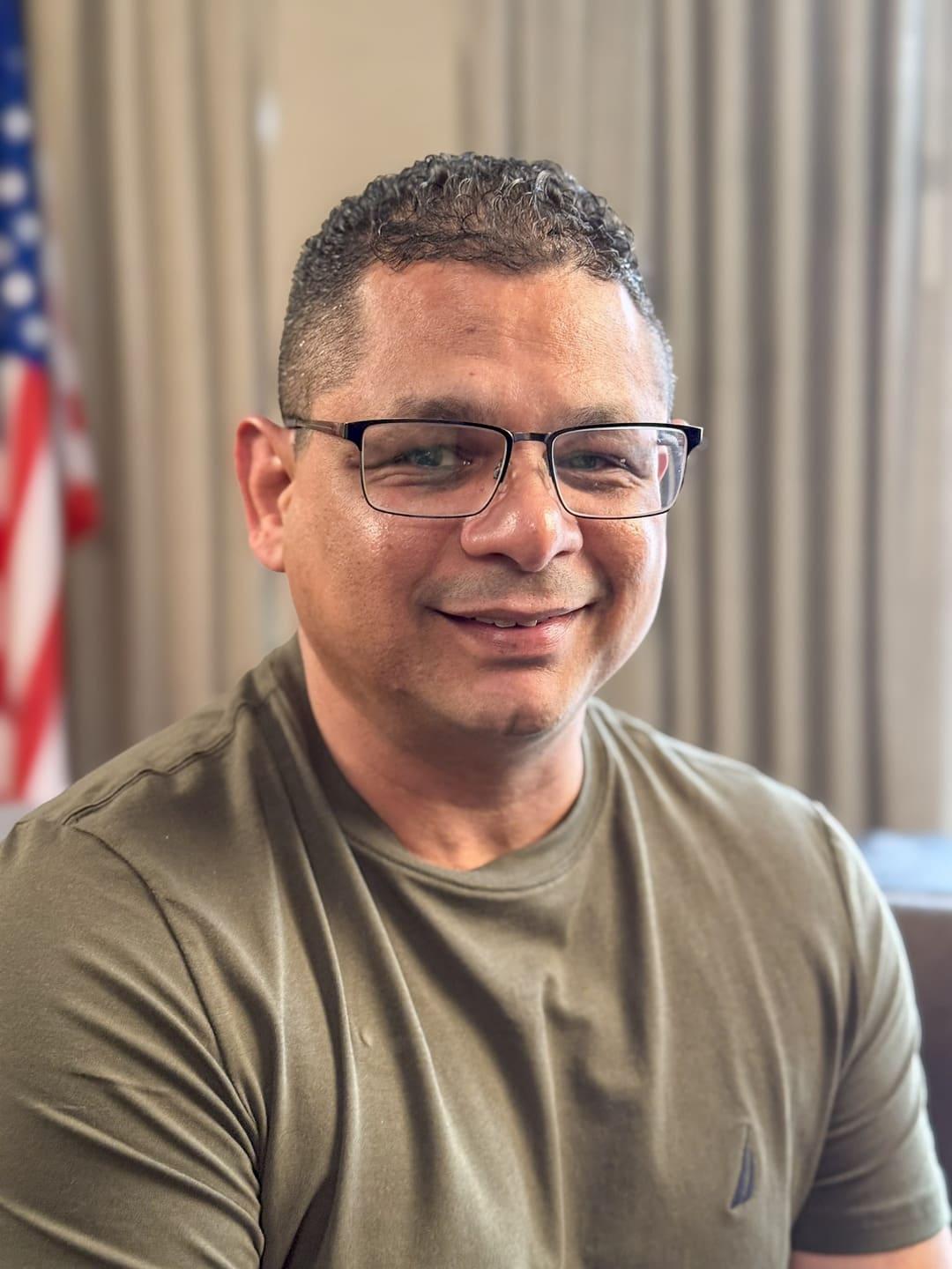 A man wearing glasses sits indoors and looks toward the camera.