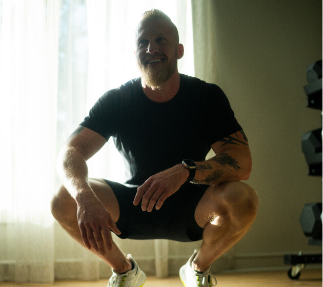 A person squatting indoors on a mat, resting after a workout.