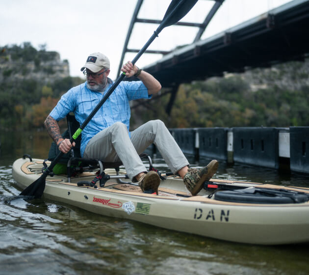 A person kayaks on a river beneath a bridge.