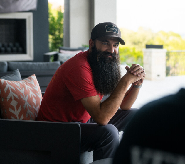 A bearded man in a red shirt sits outdoors and talks with another person in a relaxed