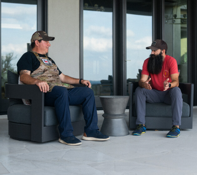 Two men sit facing each other outdoors, having a conversation.