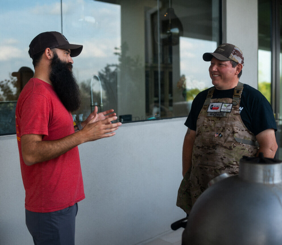 Two men stand outdoors and talk, one gesturing while the other listens.