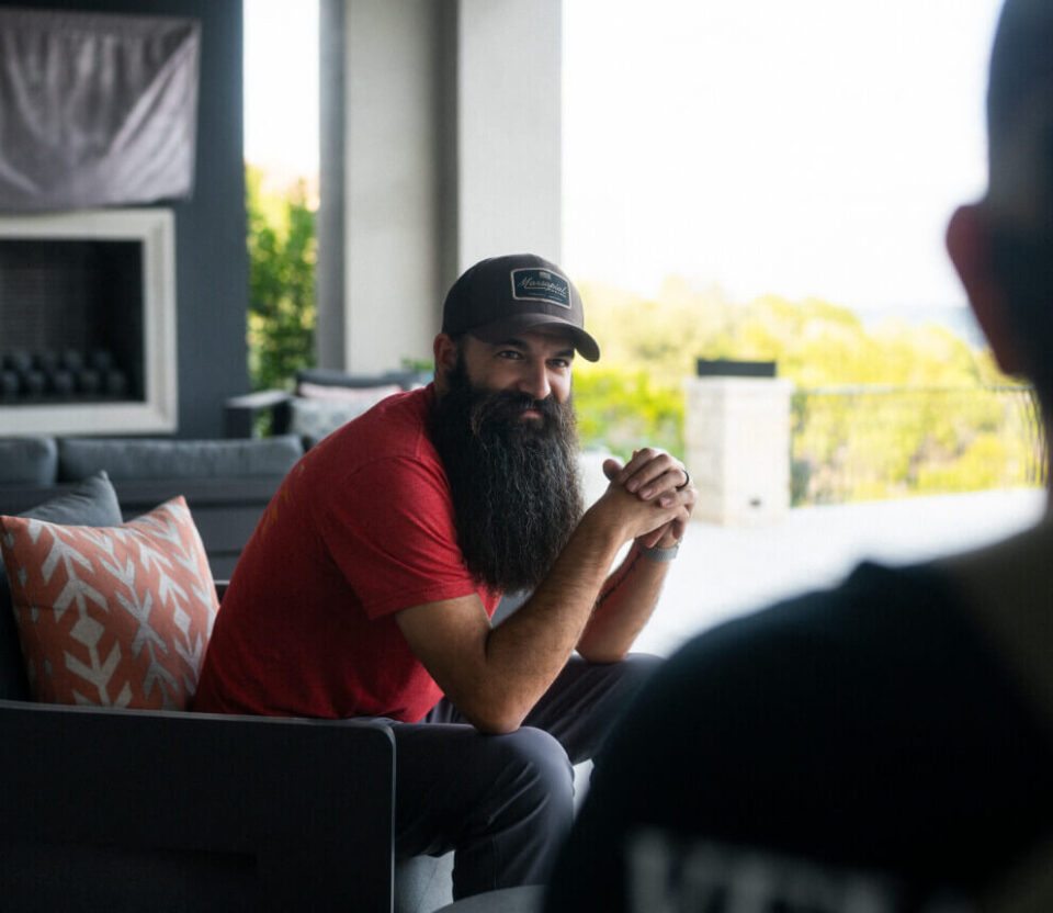 A bearded man in a red shirt sits outdoors and talks with another person in a relaxed setting.